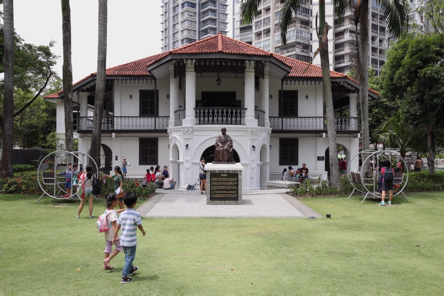 White colonial-style building with red tile roof, statue in front, people in park setting.